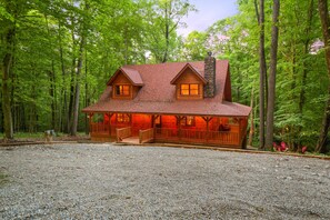 A log cabin with a porch and a chimney, surrounded by a forest.