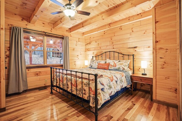 Un dormitorio de cabaña de madera con cama, ventilador de techo, ventana con cortinas y mesita de noche con lámpara.
