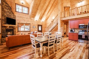 Interior de una cabaña de madera con comedor, cocina, chimenea y una escalera.