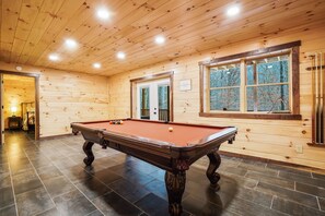 Una sala con paneles de madera, una mesa de billar, una ventana con vista a árboles y una puerta que da a otra habitación.