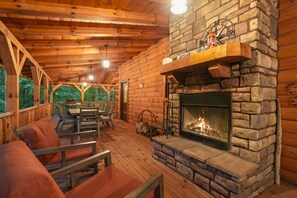 Interior de una cabaña de troncos con chimenea de piedra, vigas de madera en el techo y un área de estar al aire libre techada.