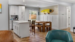 A modern kitchen with white cabinetry, a wooden dining table, and a surfboard mounted on the wall.