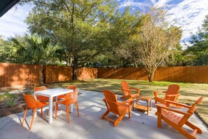 An outdoor patio with a white table and orange chairs, surrounded by a wooden fence and trees. - Cozy Home Near Folly Field Beach (Hilton Head Island)