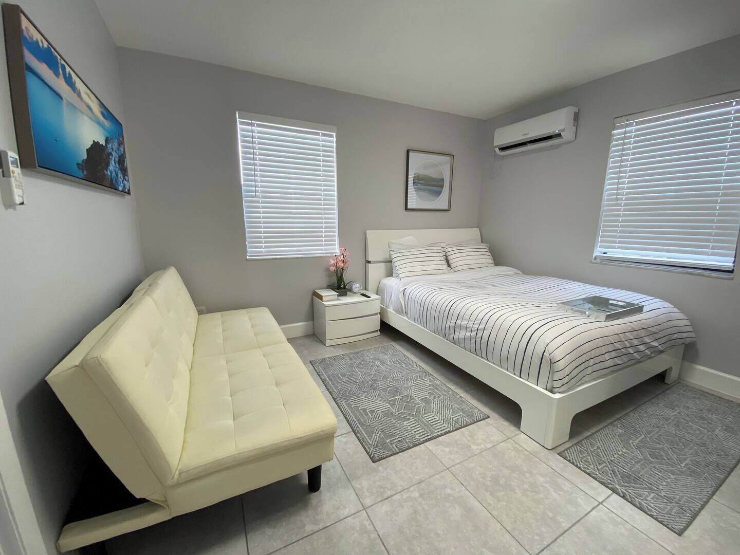 A bedroom with a bed, a sofa, a nightstand, and two windows with blinds.