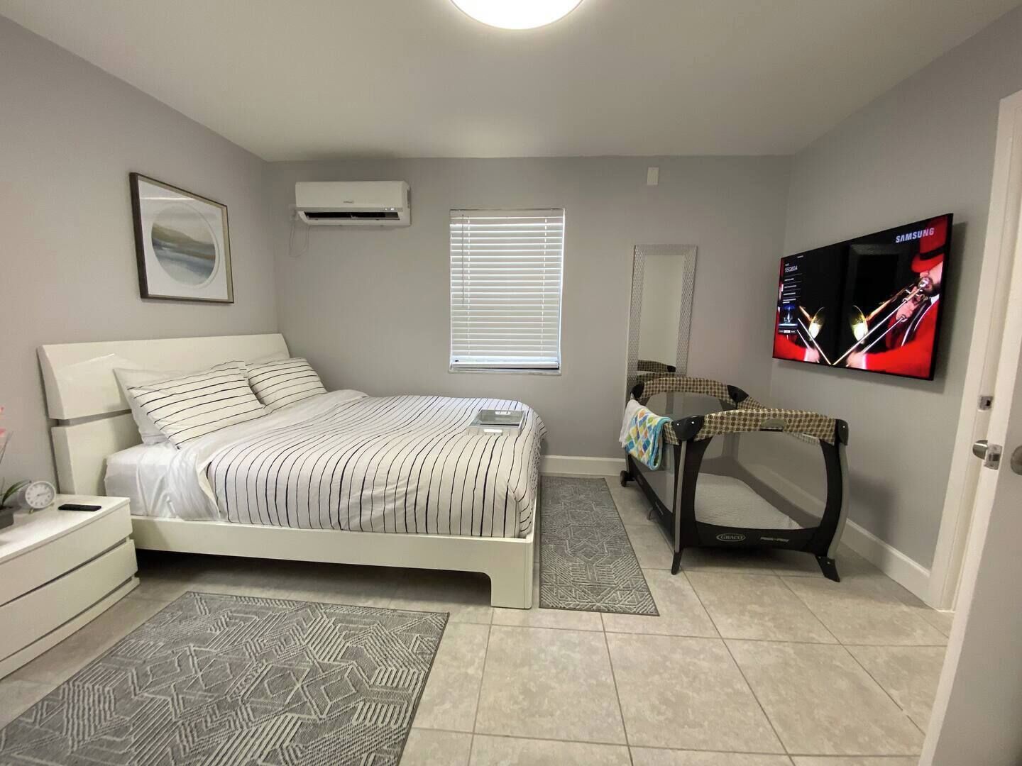 A bedroom with a bed, a TV, a mirror, and a window with blinds.