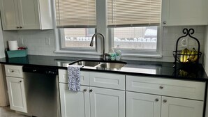 A kitchen with white cabinets, a stainless steel dishwasher, a black countertop, a sink with a faucet, and a window with blinds. - Downtown Home Away from Home 3BR/3BA (Rochester)