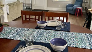 A dining area with a wooden table set for four, including plates, bowls, cutlery, and glasses. There are two chairs, a blue armchair, and a small stool. The room has a door, windows with curtains, and a lamp. - Downtown Home Away from Home 3BR/3BA (Rochester)
