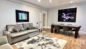 Ein modernes Wohnzimmer mit grauem Sofa, einem Glastisch, einem Essbereich mit Holztisch und Stühlen sowie einem großen, an der Wand befestigten Fernseher.