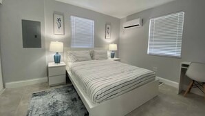Un dormitorio moderno con una cama grande, dos mesitas de noche con lámparas, aire acondicionado de pared y una ventana con persianas.
