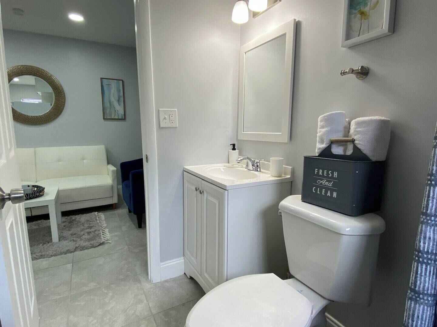 Un baño moderno con inodoro, lavamanos y espejo. En la zona de estar contigua hay un sofá blanco.