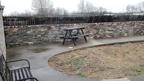 An outdoor seating area with benches and picnic tables, surrounded by a brick wall and a fence.