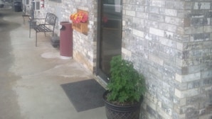 A building with a brick exterior, a door, a window, a chair, a trash can, and a potted plant.
