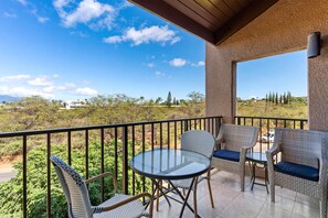 Un balcón con mobiliario de mimbre y una mesa de vidrio, con vistas a un paisaje con árboles y edificios.