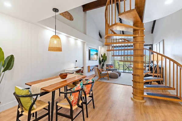 A modern interior with a wooden staircase, a dining area with a wooden table and chairs, and a large window offering a view.