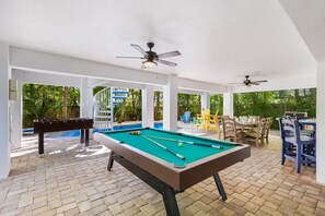 Une table de billard avec des queues et des billes, une table de baby-foot, et un espace extérieur aménagé avec des chaises et une table.