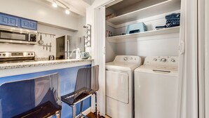 Cocina compacta con microondas, estufa y lavaplatos. Hay dos lavadoras blancas y una silla azul.