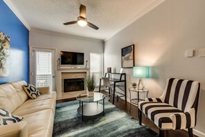 A living room with a fireplace, a ceiling fan, a TV, a sofa, a striped armchair, and a small table with a lamp.