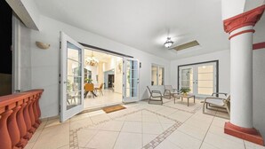 Un espacio interior amplio con suelo de baldosas, zona de comedor con sillas y mesas, y un gran ventanal que da a otra habitación.