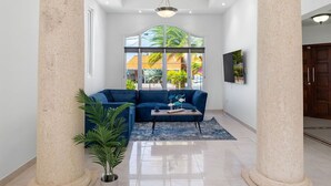 Amplia sala de estar con un sofá azul, una mesa de centro y una planta en maceta. Se observan dos columnas grandes y una lámpara de techo.