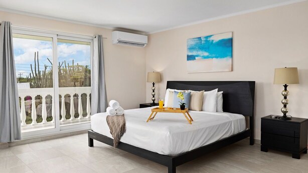 A hotel room with a bed, two bedside tables, a window with curtains, an air conditioning unit, and a painting on the wall. - ~Sleeps 12 ~Spacious ~Private Pool ~Great Location (Noord)