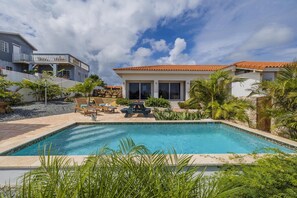 A swimming pool with lounge chairs, a patio area with a table and chairs, and a view of a house with a red-tiled roof. - Serene Modern Studio with Pool & Nature Views (Santa Cruz)