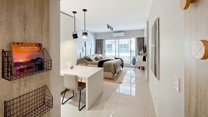 Een moderne hotelkamer met een bed, nachtkastje en een bureau met stoel. Boven het bed hangen twee hanglampen en er is een groot raam met gordijnen.