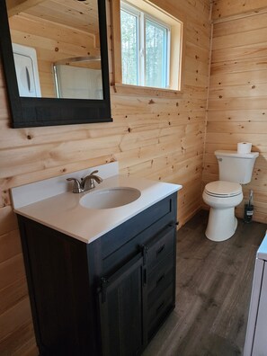 Une salle de bain rustique, avec des murs en bois, un lavabo blanc, des meubles noirs, un miroir et des toilettes.