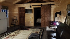 Interior acogedor de una cabaña de madera con techo de madera, televisor montado en la pared, refrigerador, sofá y alfombra estampada.