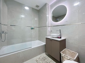 A modern bathroom with a glass-enclosed shower, a white bathtub, a round mirror with a light, and a sink with a marble countertop.