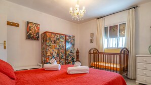 Un dormitorio con una colcha roja, una cuna, una pared con un collage de imágenes y una araña.