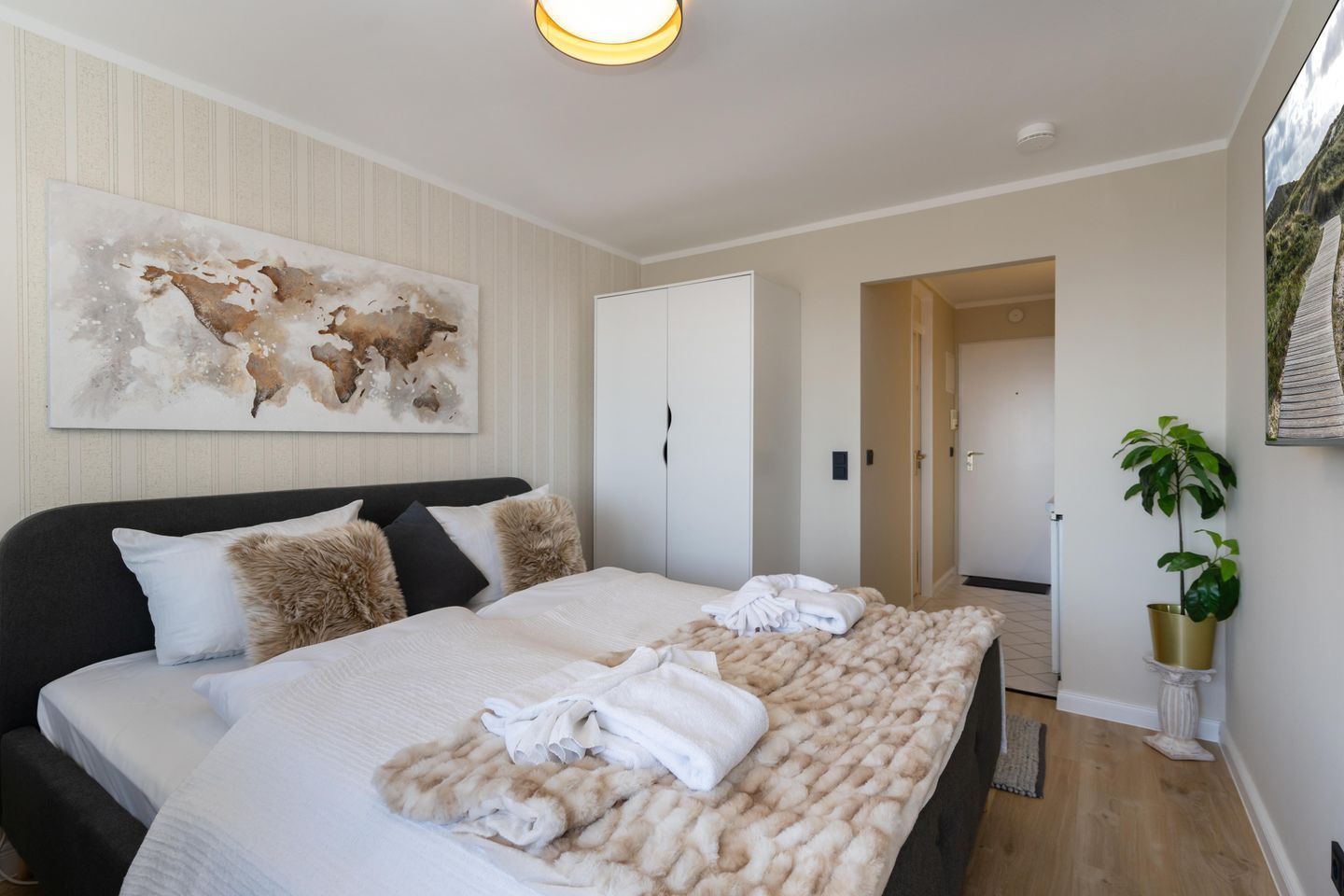 A modern hotel room with a large bed, a painting on the wall, a wardrobe, and a potted plant.