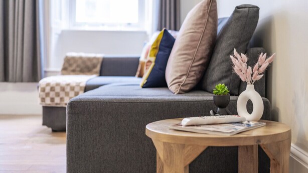 En moderne stue med en grå sofa, mange puder, et lille rundt træbord med en fjernbetjening, en vase med tørrede planter og et vindue med gardiner.