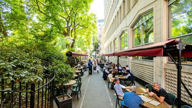 Un café al aire libre estrecho con mesas y sillas, rodeado de árboles y edificios.