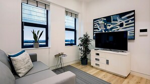 Una sala de estar moderna con un sofá gris, un televisor de pantalla plana sobre un mueble blanco y dos ventanas con vistas al mar.