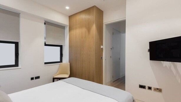 A modern bedroom with a large bed, a wooden wardrobe, a chair, and a TV mounted on the wall.