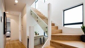 Una escalera moderna con escalones de madera y barandillas de vidrio, que conduce a un dormitorio con cama y un pequeño escritorio.