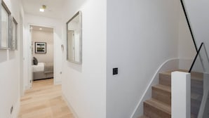 Een moderne gang met een houten vloer, een bed op de achtergrond en een trap met houten treden en een glazen leuning.