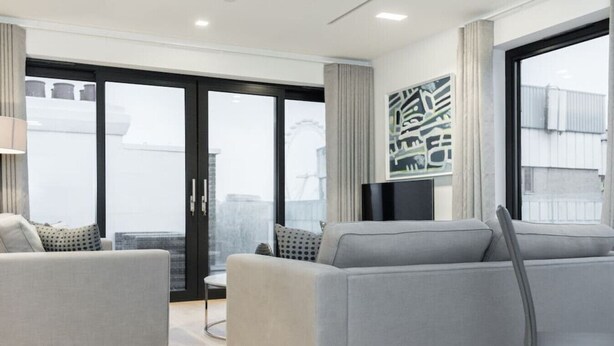 Een moderne woonkamer met een grijze zithoek, een flatscreen televisie en een groot raam met gordijnen.