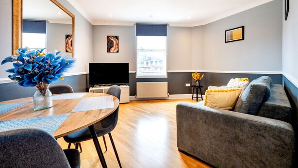 A modern living room with a grey sofa, a wooden dining table with chairs, a flat-screen TV, and artwork on the walls. - Elegant 1 Bedroom in Cambridge Circus (Greater London)
