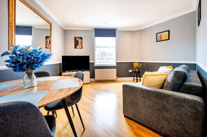 A modern living room with a grey sofa, a wooden dining table with chairs, a flat-screen TV, and artwork on the walls.