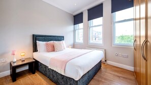 A bedroom with a large bed, a nightstand with a lamp, and a view of the outside through large windows. - Elegant 1 Bedroom in Cambridge Circus (Greater London)