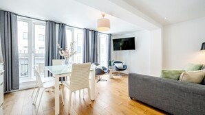 A modern living room with a grey sofa, a dining table with white chairs, and a TV mounted on the wall. - Splendid 2 Bedroom in Covent Garden (Greater London)
