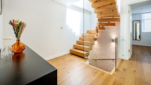 Una escalera moderna con escalones de madera y barandas de cristal en una habitación luminosa y de mobiliario sencillo.