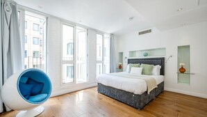 Un dormitorio moderno con una cama grande, una silla azul llamativa y ventanas amplias.