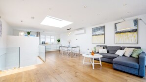 Un espacio habitable amplio con un sofá gris, una mesa de centro blanca y redonda, y una mesa de comedor de vidrio con sillas. Se observa una zona de cocina al fondo con armarios blancos y un ventanal.