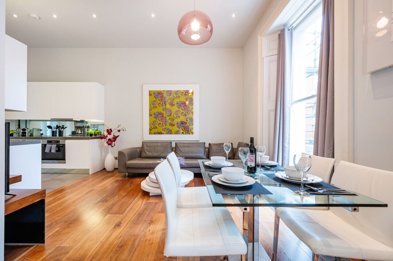 A modern living room with a dining area, white furniture, a glass table, and a kitchen in the background.