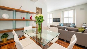 Ein modernes Wohnzimmer mit einem Esstisch aus Glas, weißen Sofas und einem Holzregal mit Dekorationen.