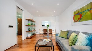 A modern living room with a glass coffee table, a grey sofa with cushions, a wooden floor, a dining area with chairs, and a wall-mounted abstract painting. - Chic 2 Bedroom in South Kensington (Greater London)
