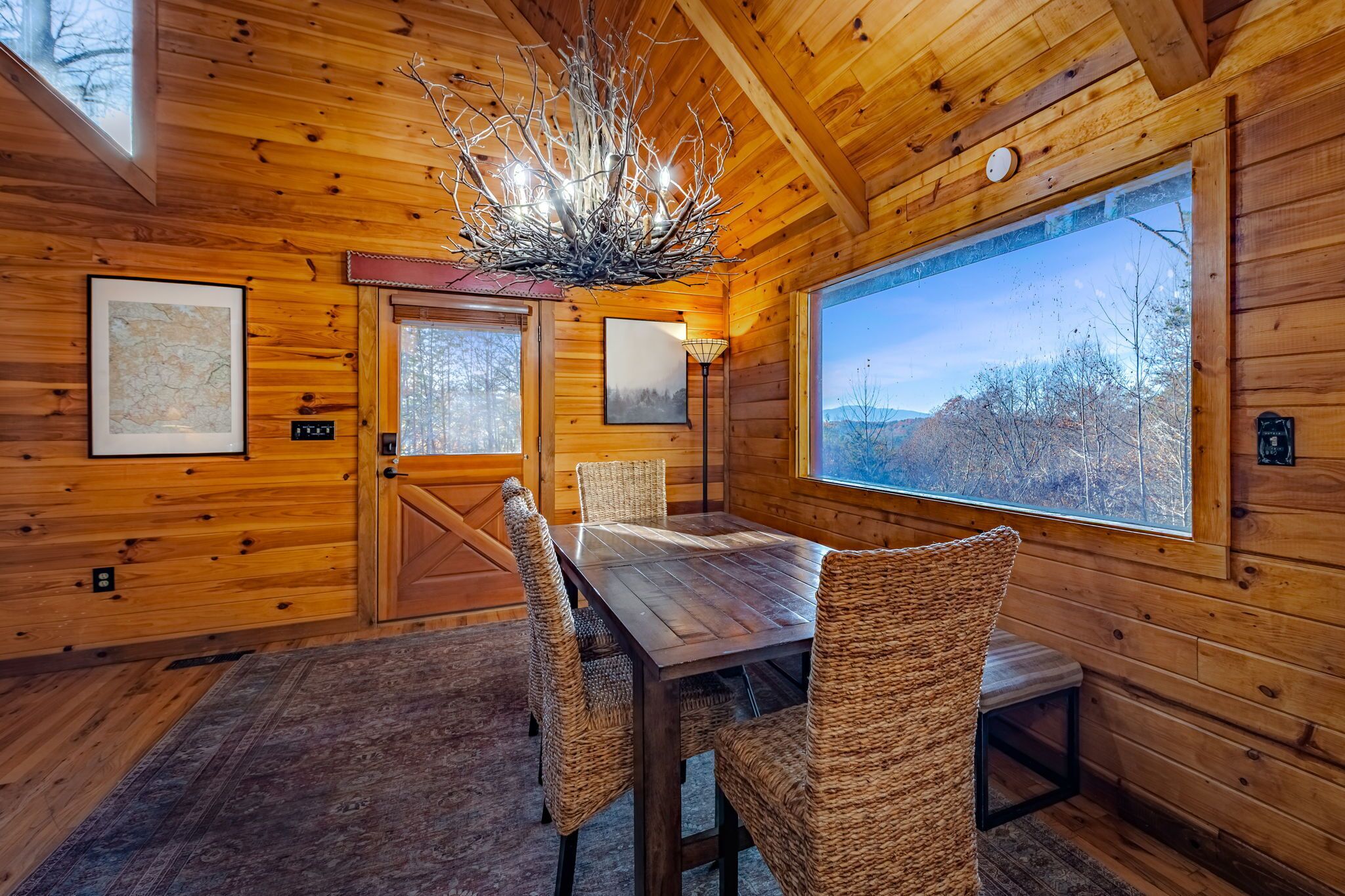 A wooden cabin interior with a dining table, wicker chairs, a chandelier, framed pictures, and large windows offering a view of the outdoors.