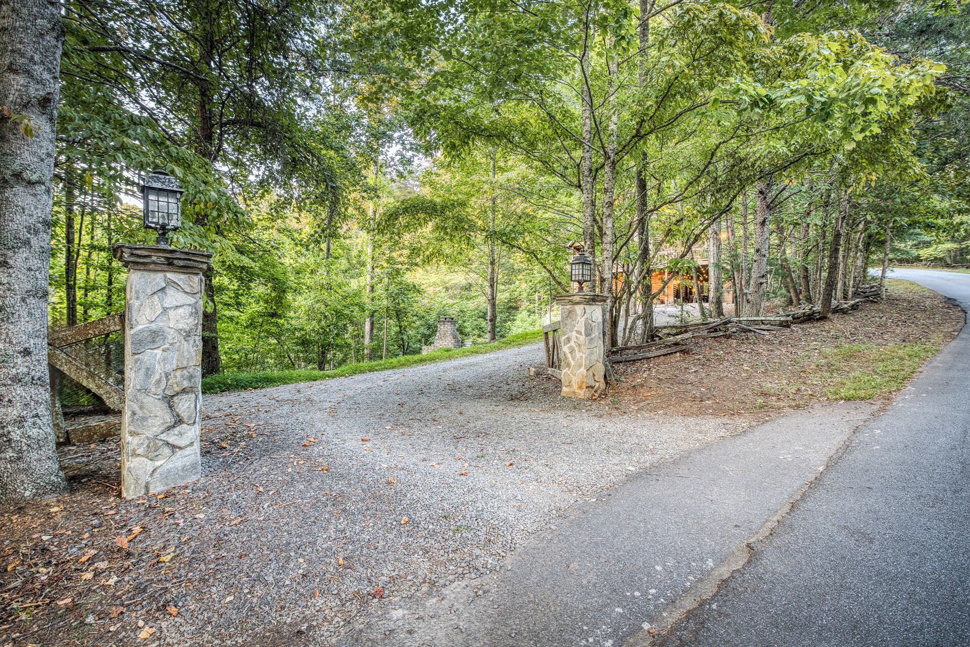 A gravel driveway leading to a stone gate with a lantern post, surrounded by trees and a building in the background.